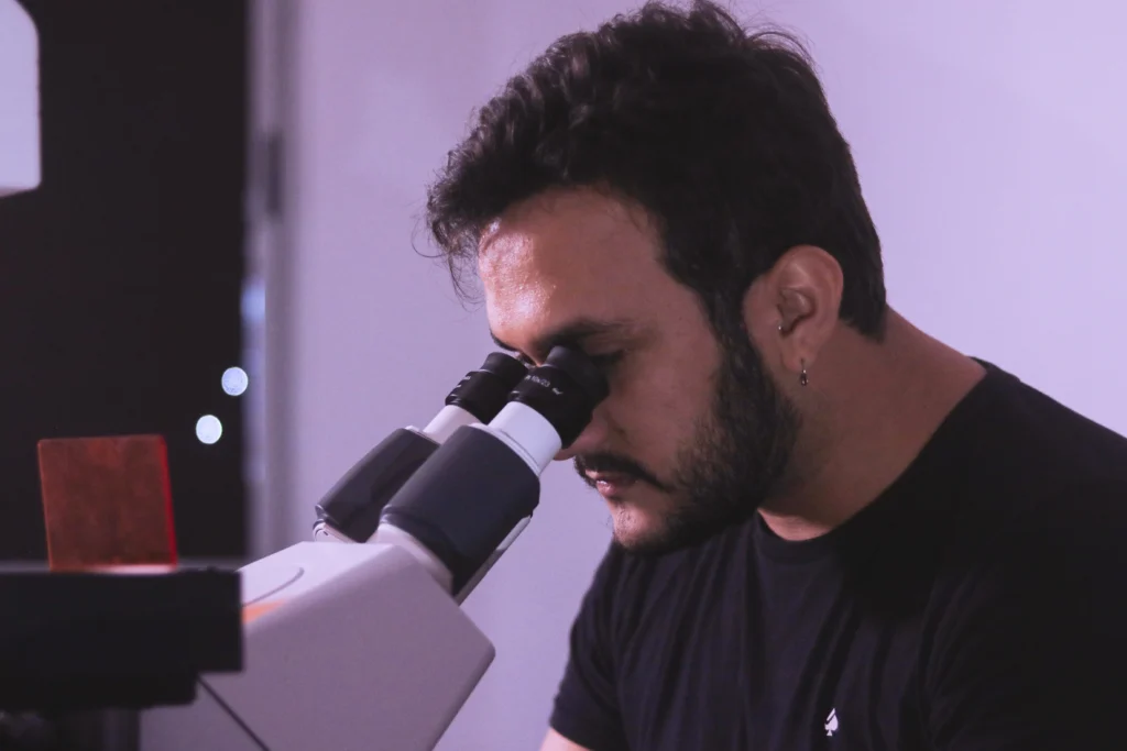 Fotografia de um jovem homem de pele clara, cabelos e barba curtos e escuros, usando camiseta preta e um brinco de argola na orelha esquerda. Ele aparece de perfil, inclinado para a frente enquanto observa através das oculares de um microscópio branco em um laboratório com iluminação suave e tom ligeiramente arroxado.