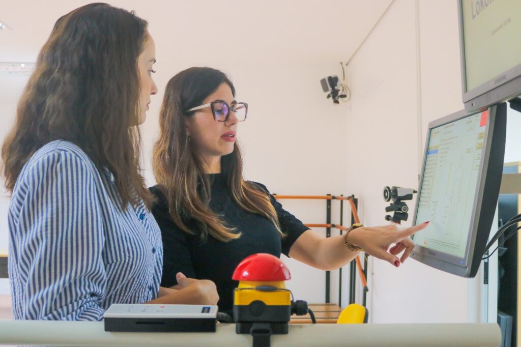 Fotografia de duas mulheres em um ambiente iluminado analisando dados juntas. A mulher à esquerda veste uma camisa listrada de azul e branco, enquanto a mulher à direita usa óculos de armação preta e blusa preta, apontando o dedo indicador para a tela de um monitor que exibe esquemas do corpo humano e parâmetros de controle. Em primeiro plano, destaca-se um botão de emergência vermelho e amarelo.