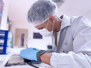 Uma foto de um profissional de laboratório, de perfil, usando jaleco branco, touca descartável e máscara branca, além de luvas azuis. Ele está com o braço levantado e parece estar manuseando ou verificando algo numa superfície vertical à esquerda (talvez um equipamento ou painel). No canto superior esquerdo há uma caixa/plástico azul com alguns compartimentos. O fundo é claro, com armários ou estruturas desfocadas, dando aparência de ambiente de laboratório ou área técnica.