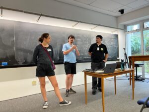 Uma foto de uma sala de aula com três pessoas em pé na frente de um quadro negro. Há uma mulher à esquerda, de cabelo preso em coque, usando blusa escura e shorts escuros, com uma bolsa tiracolo. No centro há um homem de pele clara com barba e cabelo curto, usando camiseta azul clara e shorts escuros, com o braço dobrado na frente do corpo. À direita há um homem de pele morena, cabelo curto, usando camiseta preta e calça jeans, com um crachá no peito. Na frente deles há uma mesa de madeira com um notebook aberto e outros itens pequenos em cima, além de um tripé ao lado. O quadro negro atrás tem escrita com giz, mas o texto não está legível nesta imagem.