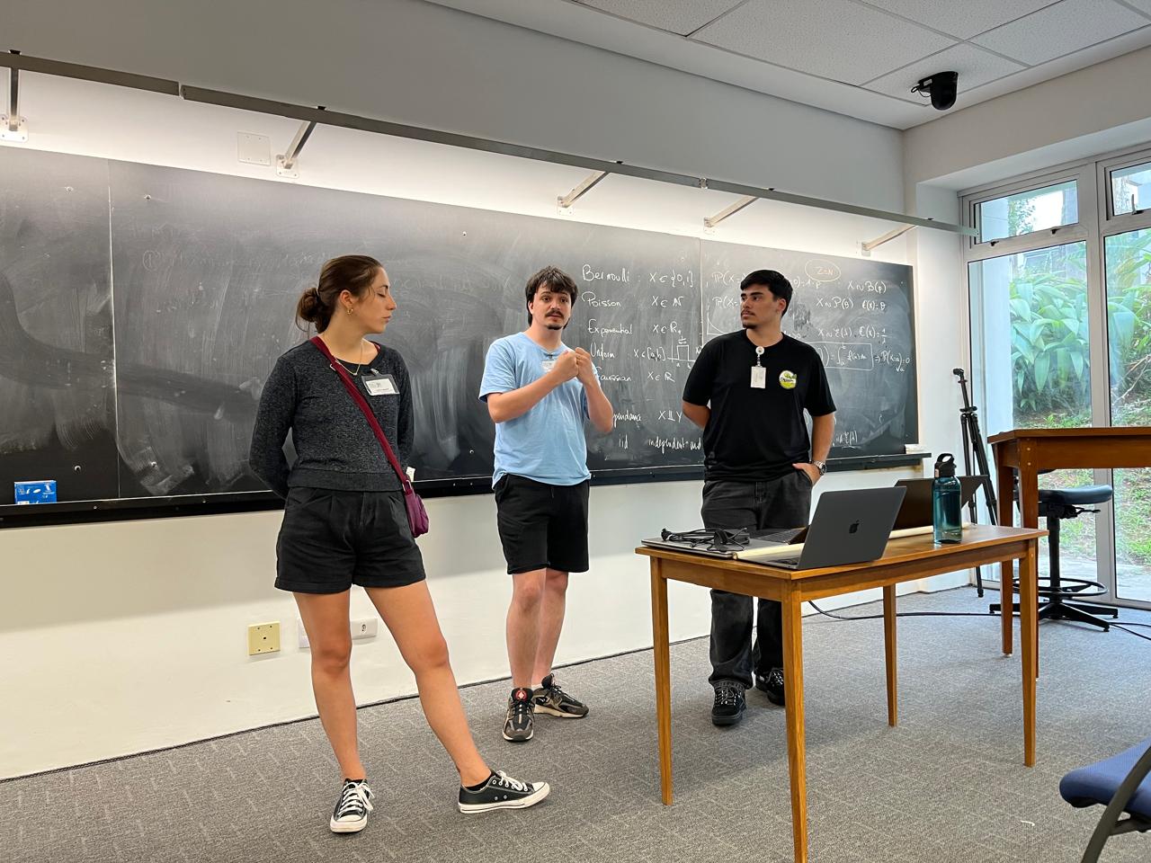 Uma foto de uma sala de aula com três pessoas em pé na frente de um quadro negro. Há uma mulher à esquerda, de cabelo preso em coque, usando blusa escura e shorts escuros, com uma bolsa tiracolo. No centro há um homem de pele clara com barba e cabelo curto, usando camiseta azul clara e shorts escuros, com o braço dobrado na frente do corpo. À direita há um homem de pele morena, cabelo curto, usando camiseta preta e calça jeans, com um crachá no peito. Na frente deles há uma mesa de madeira com um notebook aberto e outros itens pequenos em cima, além de um tripé ao lado. O quadro negro atrás tem escrita com giz, mas o texto não está legível nesta imagem.