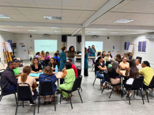 Uma foto de um grupo de pessoas sentadas em cadeiras, em uma sala grande, em atividade em grupo. Há duas fileiras principais de cadeiras formando um corredor no meio, com pessoas conversando e voltadas umas para as outras. Algumas pessoas estão em pé ao fundo, perto da parede.