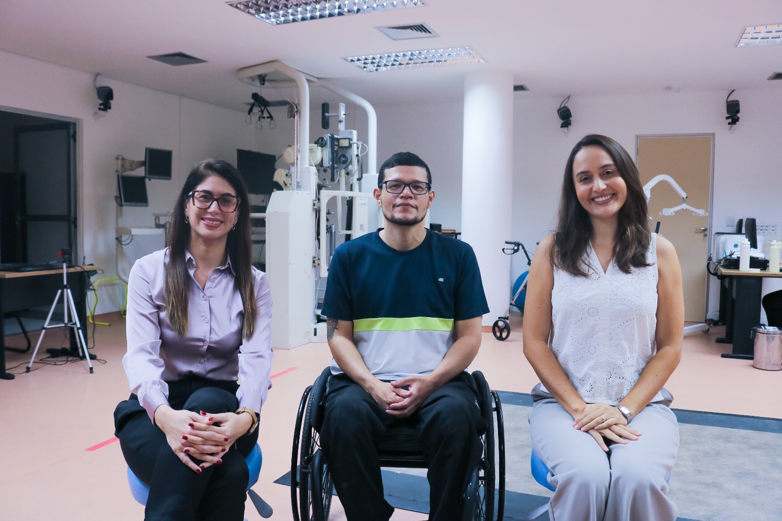 Três pessoas em um ambiente de fisioterapia. Ao centro, há um homem em cadeira de rodas, usando óculos e camiseta azul‑escura com uma faixa clara no peito. Ele está sorrindo levemente e com as mãos apoiadas no colo. À frente dele, mais perto da câmera, está uma mulher sentada num banco azul. Ela tem cabelos castanhos escuros, compridos, e está sorrindo bastante. Usa blusa branca sem mangas rendada e calça clara. As mãos estão cruzadas sobre o colo. Atrás do homem, um pouco mais distante, há outra mulher sentada. Ela também sorri, usa óculos, tem cabelos lisos castanhos, compridos, e veste camisa social lilás clara com calça escura. As mãos dela também estão cruzadas no colo. O fundo mostra vários equipamentos de clínica, mesas, monitores e aparelhos, com paredes claras e luz de teto forte.