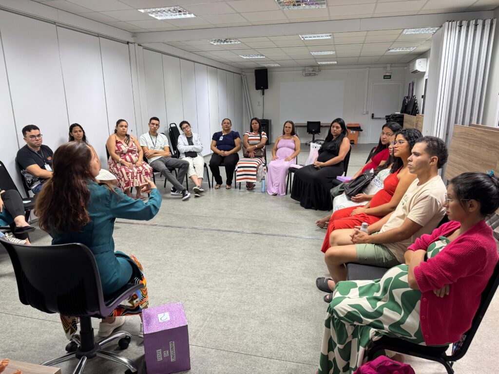 Uma sala com várias pessoas sentadas em cadeiras formando um círculo, como em uma reunião ou dinâmica de grupo. No primeiro plano, aparecem quatro pessoas sentadas em fila, de perfil, voltadas para o centro do círculo. Há uma mulher com blusa vermelha, outra pessoa com roupa clara, uma pessoa com roupa laranja e uma pessoa coberta por um tecido/roupa com estampa verde e branca.No lado oposto do círculo, perto da parede, há outras pessoas sentadas, incluindo homens e mulheres. Uma mulher com vestido listrado em tons de vermelho/rosa está com a mão no rosto. Um homem de camisa clara e calça cinza está sentado com as pernas cruzadas. Há também uma mulher de vestido claro (lilás) e uma mulher com roupa escura.O ambiente parece uma sala de treinamento ou sala de aula, com paredes claras e iluminação de teto. À esquerda há uma caixa roxa com um rótulo que diz “VESTE DA LINGUINHA”.