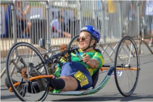 Ciclista usando capacete azul e óculos escuros, montado em uma bicicleta de estrada. A camisa é verde e amarela e o short é azul. Há uma placa amarela com o número “302” presa no lado do quadril. A bicicleta tem guidão curvado e rodas finas; aparece uma roda de outra bicicleta no canto inferior. Ao fundo, há grades metálicas e algumas pessoas desfocadas atrás delas.