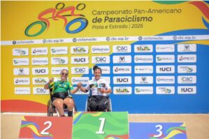 Duas mulheres em cadeiras de rodas em um pódio, em frente a um painel de patrocinadores. À esquerda, no lugar marcado com o número 2, uma pessoa com uniforme verde e amarelo (parece uma camisa de ciclismo) e shorts preto, usando óculos escuros, segurando um objeto claro na mão. No centro, no lugar marcado com o número 1, uma pessoa com camisa branca e calça escura, com uma medalha no pescoço, segurando um objeto na mão (parece um bichinho de pelúcia ou mascote). O pódio tem as faixas coloridas e os números 1, 2 e 3 (o 3 aparece no canto inferior esquerdo da imagem). No painel de fundo aparecem vários logotipos e textos repetidos, incluindo “UCI” e “CBC”. No lado direito, em um fundo amarelo, dá para ler “Campeonato Pan-Americano de Paraciclismo Pista e Estrada / 2026”.