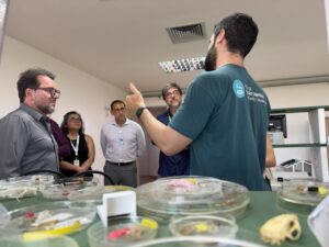 Membros da CAACG e da diretoria do ISD visitando um laboratório de neuroengenharia. Na foto, cinco pessoas aparecem ao redor de uma bancada do laboratório. Em primeiro plano, de costas, há um homem com cabelo e barba escuros usando uma camiseta verde-azulada; no ombro da camiseta há um logotipo e texto branco (não está totalmente legível). O braço direito dele está levantado, com a mão apontando para cima, como se estivesse falando ou explicando algo. Do outro lado da mesa, à esquerda, há um homem de meia-idade com pele clara, cabelo escuro com um pouco de grisalho, barba grisalha curta e óculos, usando uma camisa social azul-clara; ele está de perfil, voltado para o grupo. Atrás dele aparece uma mulher com pele clara, cabelo escuro e óculos, usando uma blusa roxa e um crachá. Mais ao centro, há um homem com pele clara, cabelo curto escuro e óculos, usando camisa social clara e calça cinza; ele está de frente para a câmera. À direita dele, há uma pessoa com pele clara e cabelo curto grisalho, usando óculos e uma camisa escura, sentada e voltada para o homem em primeiro plano. No fundo, a parede é bege, com uma luminária retangular no teto e uma grade de ventilação. Também há uma estante ou móvel com equipamento eletrônico ao fundo.