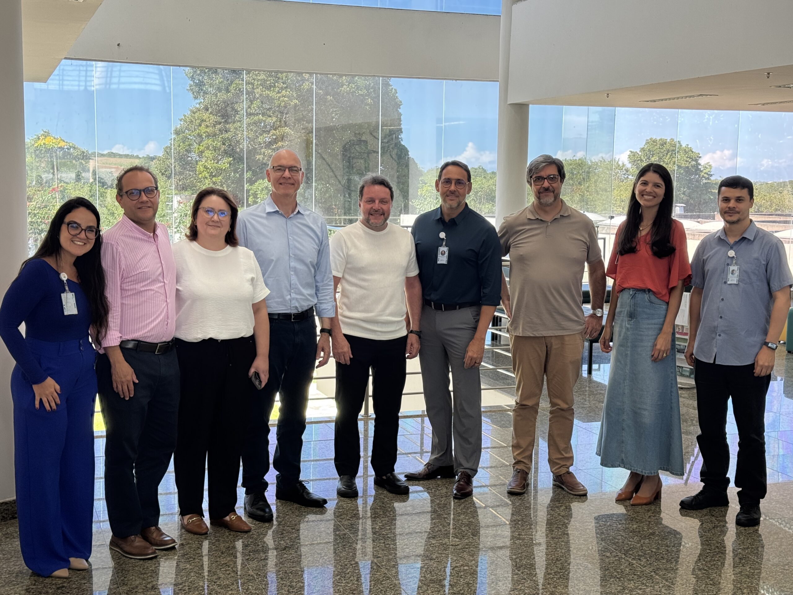 Grupo de pessoas posando para foto em um ambiente bem iluminado, com paredes brancas e uma grande janela de vidro ao fundo.