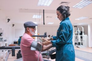 Dois adultos em um ambiente interno que parece uma clínica ou laboratório. Um homem de pele clara, usando boné cinza, óculos e camiseta rosa, está inclinado para a frente com um manguito de pressão arterial cinza no braço esquerdo. À frente dele, uma mulher de pele clara com cabelo escuro preso em um coque, usando um jaleco azul, segura um dispositivo retangular claro (parece um aparelho/monitor) com as duas mãos na altura do peito, voltado para o homem. Ao fundo aparecem equipamentos e móveis claros, com iluminação de teto em painéis retangulares.