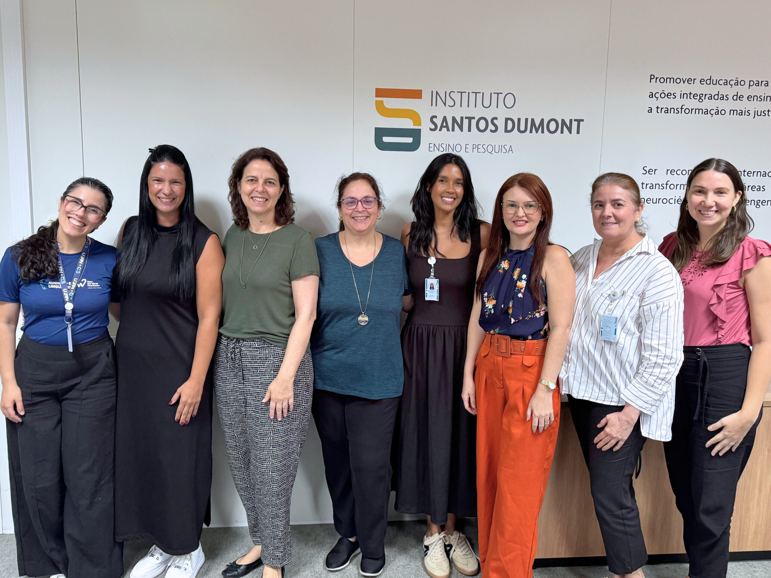 Oito mulheres de diferentes faixas etárias estão de pé, enfileiradas, olhando para a câmera. Elas estão em uma sala com a logomarca do Instituto Santos Dumont em uma parede atrás delas. Elas sorriem para a foto.