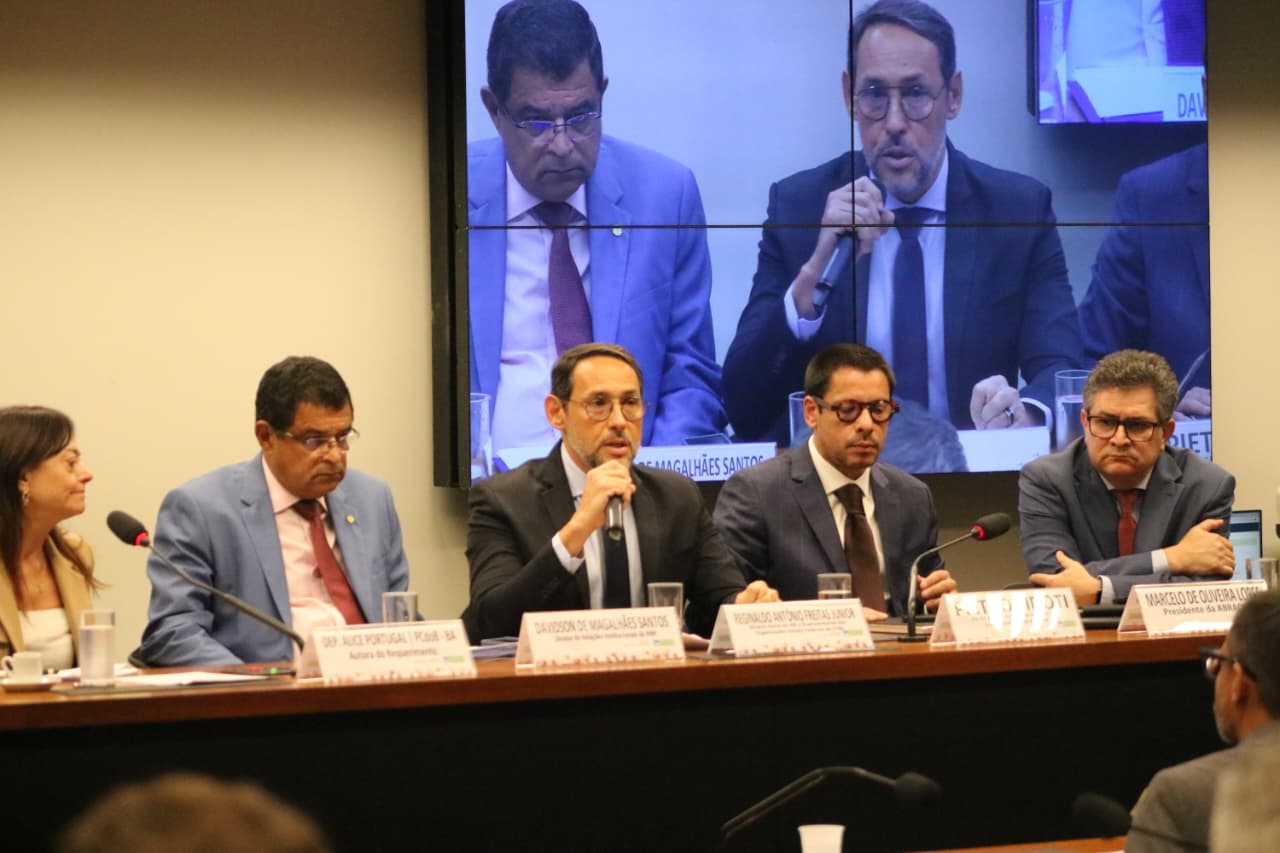 Uma mesa de reunião em audiência formal, com vários homens de terno sentados lado a lado atrás de um balcão de madeira. Eles usam ternos em tons de cinza ou azul, com gravatas, e estão em ambiente de aparência oficial. Alguns estão falando em microfones, outros parecem estar ouvindo ou anotando. Atrás deles há um grande painel de vídeo azul, dividido em quadrantes, exibindo em close o rosto de um dos homens enquanto fala ao microfone, repetido ou enquadrado de maneiras ligeiramente diferentes. A iluminação é clara e o clima é de sessão séria, como numa audiência pública ou reunião de comissão.