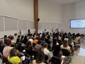 Auditório com muitas pessoas sentadas em cadeiras, voltadas para a frente, em um evento que parece ser uma palestra ou treinamento. A imagem está girada de lado. Há um corredor central e fileiras cheias; várias pessoas estão escrevendo em cadernos ou usando celular. Predominam adultos, com roupas casuais (blusas, camisas, jaquetas) e cabelos de vários comprimentos; há diversidade de tons de pele. Na parte da frente, à direita, há uma tela de projeção com um slide (o texto do slide está pequeno e não dá para ler com clareza). Perto do centro da frente há uma pessoa em pé falando, com microfone, e outras pessoas próximas. O ambiente tem paredes claras, janelas grandes com persianas e uma coluna de madeira no meio.