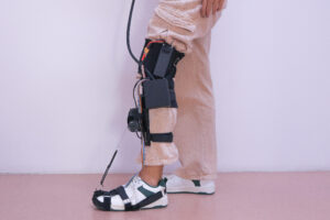 Uma perna humana estendida na horizontal com uma órtese/exoesqueleto preso do pé até a coxa. A pele visível é clara. O pé está dentro de uma bota branca e preta com várias tiras pretas. Ao longo da canela e do joelho há uma estrutura rígida preta com placas e cintas de velcro pretas prendendo a perna. Perto da região do joelho há um componente circular preto que parece uma articulação/motor, com fios coloridos passando ao longo da perna. Na parte da coxa há uma peça preta maior com uma alça e uma fivela, e um cabo grosso preto sai dessa área.