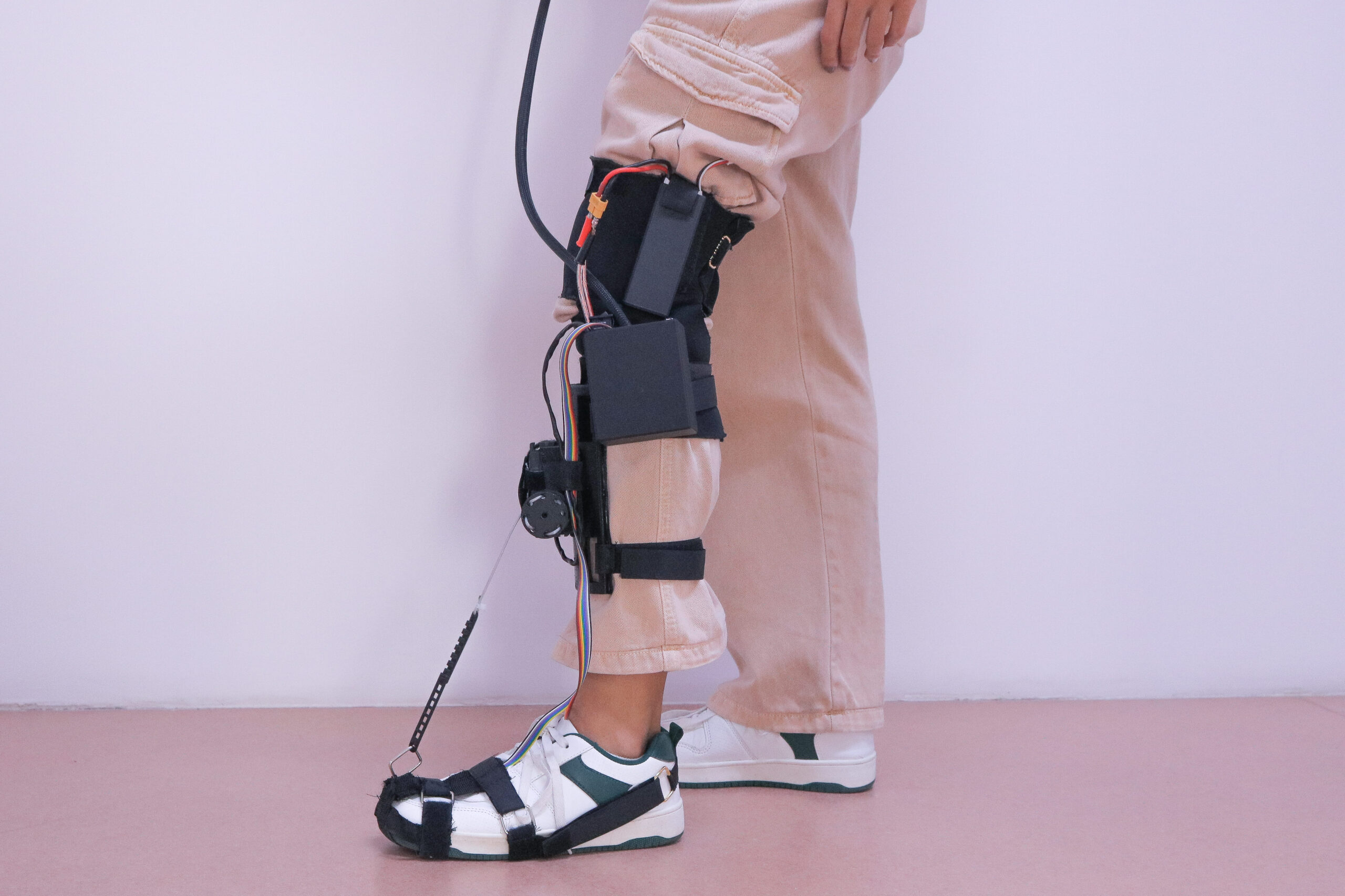 Uma perna humana estendida na horizontal com uma órtese/exoesqueleto preso do pé até a coxa. A pele visível é clara. O pé está dentro de uma bota branca e preta com várias tiras pretas. Ao longo da canela e do joelho há uma estrutura rígida preta com placas e cintas de velcro pretas prendendo a perna. Perto da região do joelho há um componente circular preto que parece uma articulação/motor, com fios coloridos passando ao longo da perna. Na parte da coxa há uma peça preta maior com uma alça e uma fivela, e um cabo grosso preto sai dessa área.