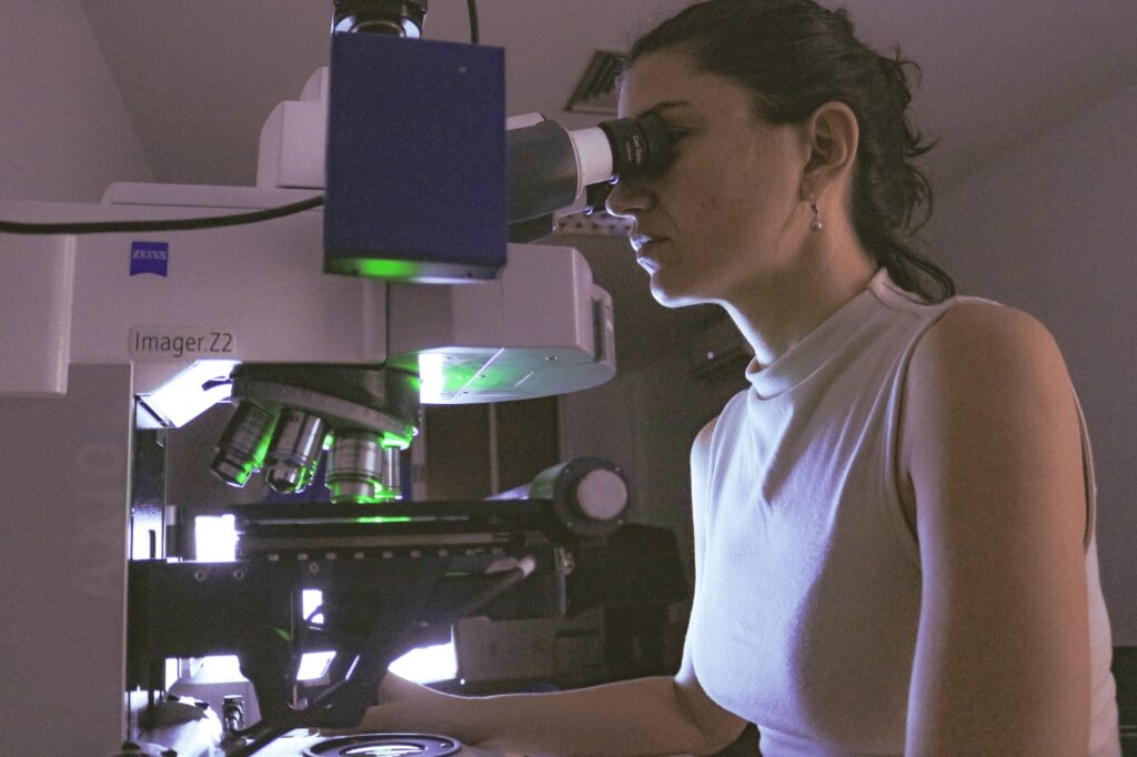 Uma mulher usando um microscópio em um laboratório. Ela está em posição lateral, com o rosto encostado na ocular do microscópio, e usa óculos e um brinco discreto. O cabelo está preso para trás em um rabo de cavalo ou coque simples. Ela veste uma blusa sem mangas de gola alta, de tecido liso e cor clara. O microscópio é grande, de bancada, com várias objetivas metálicas voltadas para baixo, iluminação interna em tom esverdeado e uma parte superior branca com a inscrição “imager Z2” e a marca “Zeiss”. Há uma caixa ou módulo retangular escuro conectado à parte superior do microscópio, possivelmente um acessório de câmera ou sistema de captura de imagem. À esquerda aparecem partes da estrutura do microscópio, com botões e rodas de ajuste visíveis.