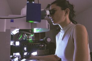 Uma mulher usando um microscópio em um laboratório. Ela está em posição lateral, com o rosto encostado na ocular do microscópio, e usa óculos e um brinco discreto. O cabelo está preso para trás em um rabo de cavalo ou coque simples. Ela veste uma blusa sem mangas de gola alta, de tecido liso e cor clara. O microscópio é grande, de bancada, com várias objetivas metálicas voltadas para baixo, iluminação interna em tom esverdeado e uma parte superior branca com a inscrição “imager Z2” e a marca “Zeiss”. Há uma caixa ou módulo retangular escuro conectado à parte superior do microscópio, possivelmente um acessório de câmera ou sistema de captura de imagem. À esquerda aparecem partes da estrutura do microscópio, com botões e rodas de ajuste visíveis.