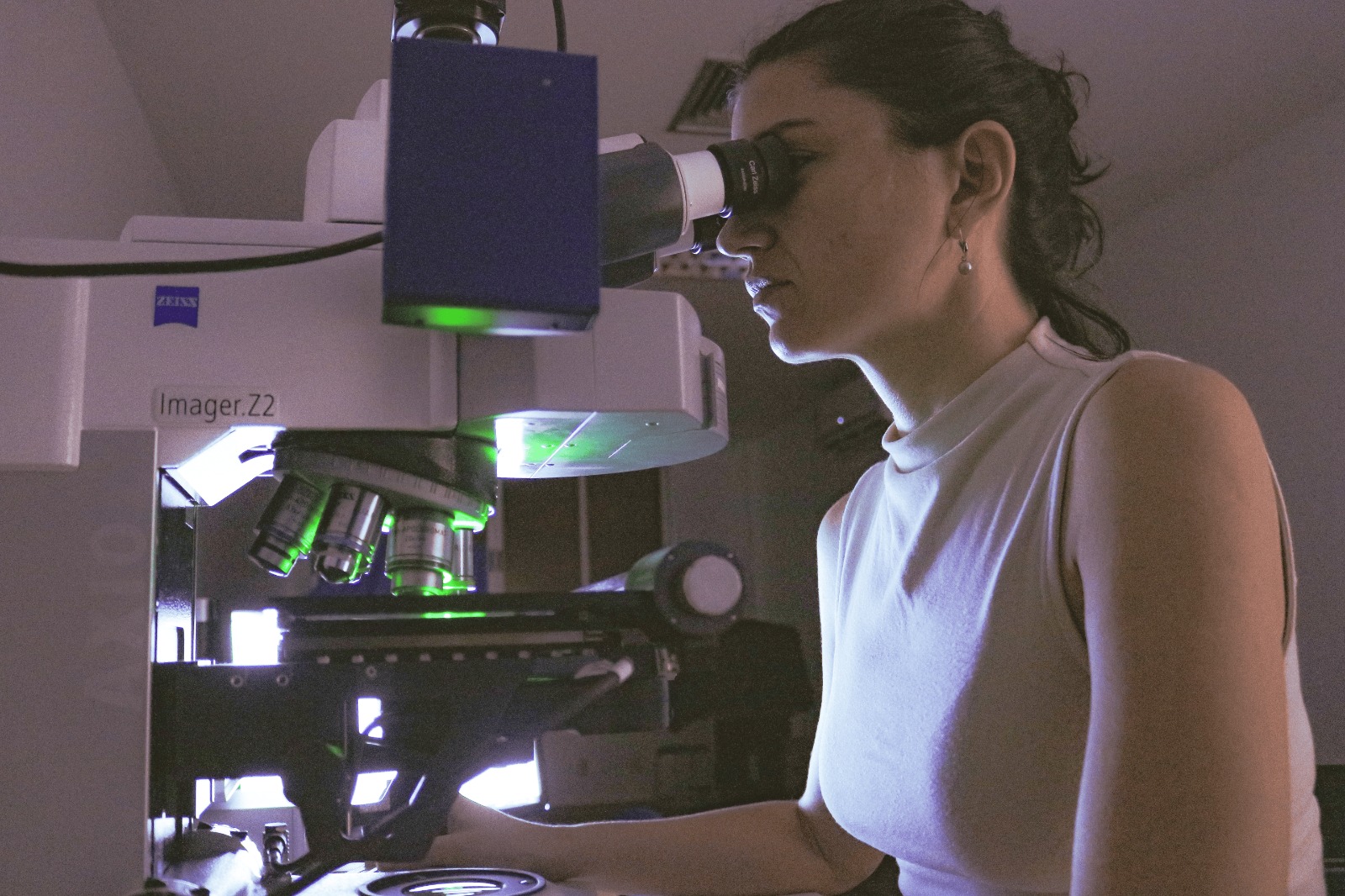 Uma mulher usando um microscópio em um laboratório. Ela está em posição lateral, com o rosto encostado na ocular do microscópio, e usa óculos e um brinco discreto. O cabelo está preso para trás em um rabo de cavalo ou coque simples. Ela veste uma blusa sem mangas de gola alta, de tecido liso e cor clara. O microscópio é grande, de bancada, com várias objetivas metálicas voltadas para baixo, iluminação interna em tom esverdeado e uma parte superior branca com a inscrição “imager Z2” e a marca “Zeiss”. Há uma caixa ou módulo retangular escuro conectado à parte superior do microscópio, possivelmente um acessório de câmera ou sistema de captura de imagem. À esquerda aparecem partes da estrutura do microscópio, com botões e rodas de ajuste visíveis.