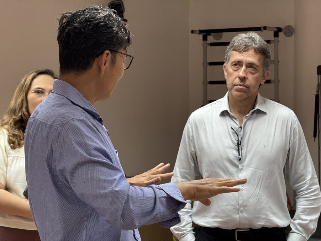 Fotografia em plano médio de três pessoas em um ambiente interno. Em primeiro plano, à esquerda e de perfil, o gerente do IIN-ELS, Edgard Morya, gesticula com as mãos enquanto explica algo. Ele usa óculos e uma camisa social azul claro. À direita, de frente para ele, o coordenador-geral do MCTI, Guilherme de Paula, ouve atentamente; ele tem cabelos grisalhos e usa uma camisa social branca. Ao fundo, à esquerda, uma mulher observa a conversa. Ao fundo, à direita, vê-se um equipamento de fisioterapia (espaldar) fixado à parede.