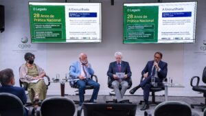 Fotografia horizontal em plano médio de uma mesa-redonda realizada em um auditório. Quatro palestrantes estão sentados em poltronas pretas giratórias, dispostas lado a lado atrás de pequenas mesas de vidro com copos de água. Da esquerda para a direita, a primeira palestrante é uma mulher de cabelos cacheados e grisalhos, usando máscara preta, colar de pedras e vestido verde-oliva com uma echarpe clara. O segundo é um homem idoso, calvo com barba branca, vestindo paletó azul-claro, camisa branca, gravata colorida e calça azul-escura. O terceiro é um homem idoso de cabelos brancos e óculos, vestindo paletó azul-escuro, camisa social clara, gravata vermelha e calça cinza, enquanto lê um papel em suas mãos. O quarto palestrante é Reginaldo Freitas Júnior, homem de pele clara, cabelos e barba aparados e escuros, vestindo terno azul-escuro e camisa clara; ele está com as pernas cruzadas e fala ao microfone. À extrema esquerda, em primeiro plano, vê-se parte das costas e da cabeça de um homem que assiste ao debate. Ao fundo, a parede branca exibe discretamente a marca do CGEE e ampara duas grandes telas de TV que transmitem o mesmo slide de uma apresentação. O slide, dividido em verde e branco, traz os títulos "O Legado - 28 Anos de Prática Nacional" e "A Encruzilhada", com logotipos da ABRACSC, IBROSS e FOSF na parte inferior.