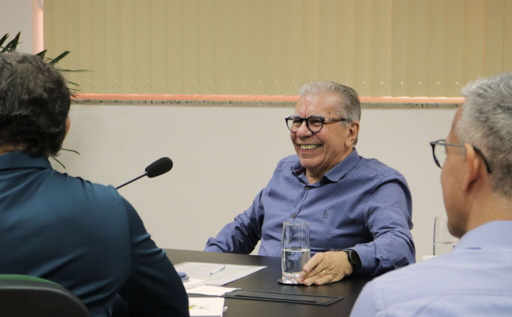 Fotografia colorida em plano médio que captura uma reunião de trabalho em torno de uma mesa escura. No centro do enquadramento, um homem de pele clara e cabelos curtos e grisalhos aparece sorrindo amplamente. Ele usa óculos de grau com armação preta e veste uma camisa social de mangas longas azul com uma textura sutil em xadrez miúdo. Ele está sentado, com o corpo voltado para a esquerda, apoiando a mão esquerda na mesa, próximo a um copo de água cheio. No pulso esquerdo, usa um relógio inteligente de pulseira escura. Em primeiro plano, as silhuetas de outras duas pessoas aparecem de costas para a câmera, emoldurando a cena: à esquerda, um homem com camisa escura de onde se projeta um microfone de mesa flexível; à direita, a nuca e o ombro de um homem vestindo camisa azul-clara. O fundo ao fundo traz uma parede clara com persianas verticais em tom bege.