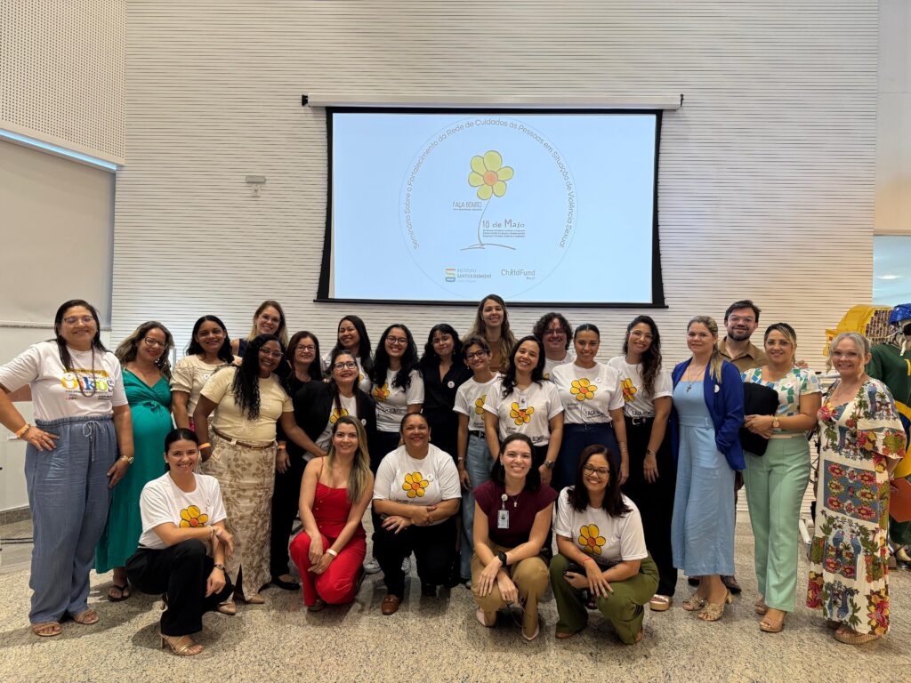 Fotografia grupal de plano inteiro com cerca de vinte e cinco participantes do seminário reunidos à frente do telão no auditório. A maioria das pessoas veste camisas brancas com o símbolo da flor amarela da campanha "Faça Bonito" ou roupas nas cores verde, azul e vermelho. Eles sorriem para a foto, divididos entre pessoas em pé e algumas agachadas na primeira fileira. Ao fundo, a tela exibe o slide principal do evento.