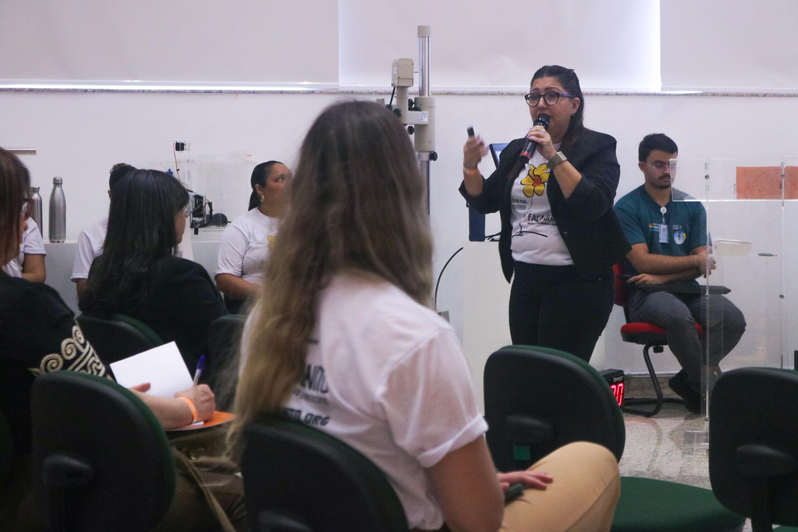 Fotografia em plano médio capturada a partir da perspectiva da plateia. No centro da imagem, uma mulher usa óculos, blazer preto e camiseta da campanha enquanto gesticula e fala ao microfone. Em primeiro plano, a silhueta desfocada de uma participante de costas preenche a parte inferior esquerda. Ao fundo, outras pessoas assistem sentadas na lateral do espaço.