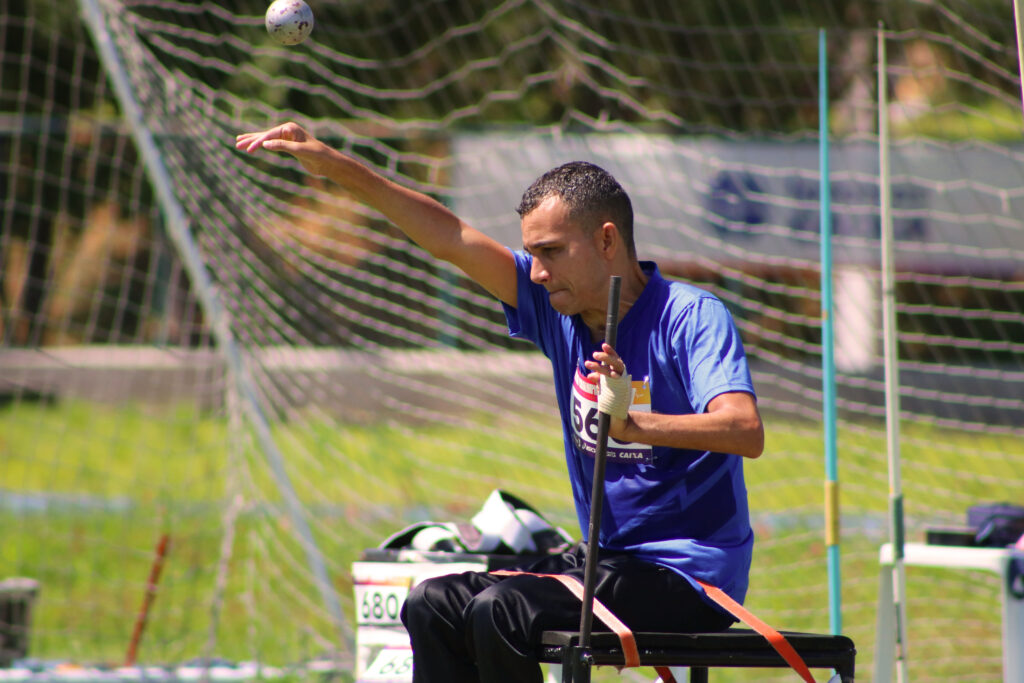 Atleta masculino sentado em uma cadeira de arremesso adaptada, fixada com tiras, realiza o movimento de arremesso de peso. Ele veste uma camiseta azul e está com o braço direito estendido para o alto, momentos após lançar a esfera de metal, que aparece no ar no canto superior esquerdo. Ao fundo, há uma rede de proteção esportiva e uma área gramada.