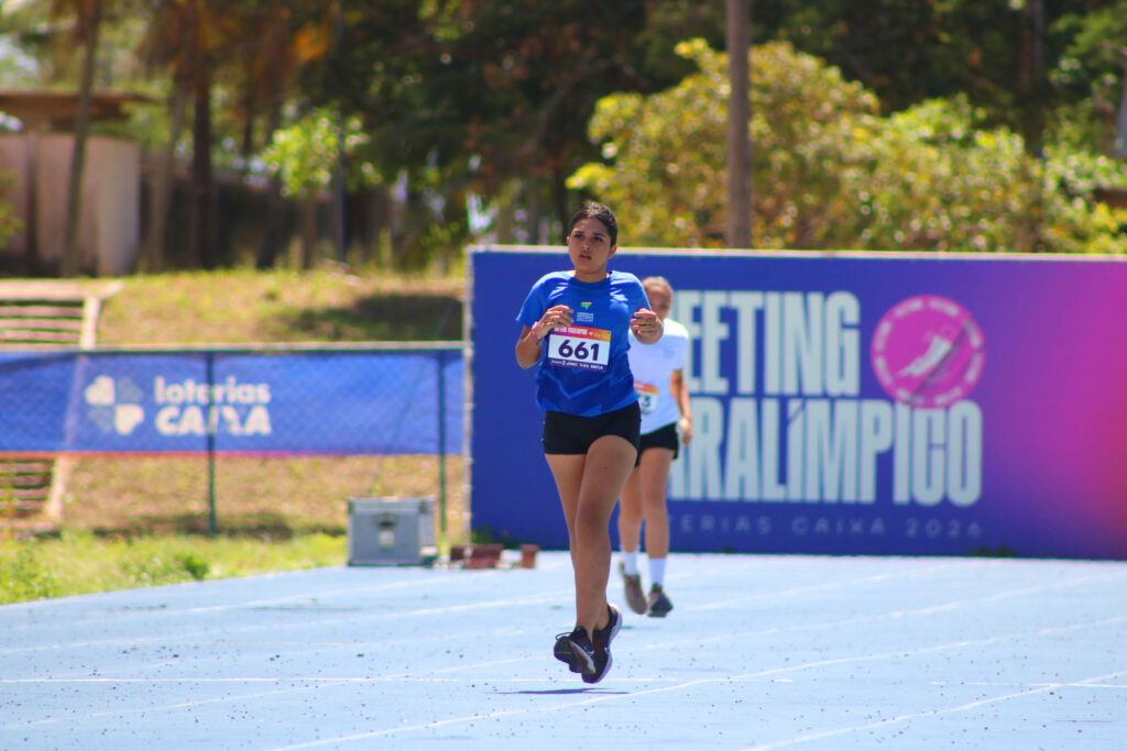 Uma jovem atleta com cabelos presos e camiseta azul corre em uma pista de atletismo azul clara durante uma competição diurna. Ela usa o número de inscrição 661 no peito. Ao fundo, outra atleta corre mais atrás e há uma grande placa roxa com a inscrição "Meeting Paralímpico Loterias Caixa 2026". A paisagem ao fundo inclui árvores e gramado sob luz solar forte.
