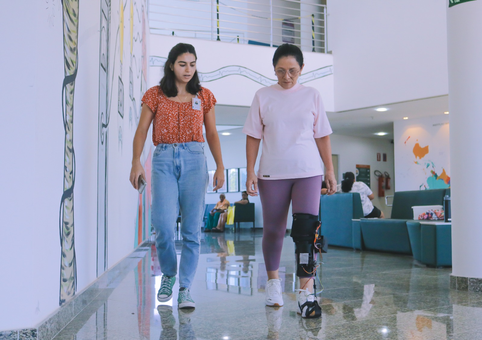Duas pessoas caminhando em um ambiente interno claro, como um saguão ou recepção. A pessoa em primeiro plano está mais à direita da imagem, tem pele clara, cabelo escuro preso para trás, usa óculos, uma blusa branca, uma bermuda ou calça justa lilás e tênis brancos. Na perna direita, logo abaixo do joelho, há uma órtese ou aparelho com estrutura metálica e partes pretas, aparentemente para apoio ou reabilitação, com alguns fios ou componentes eletrônicos visíveis. À esquerda, um pouco mais atrás, há outra pessoa com pele clara, cabelo escuro liso na altura dos ombros, usando uma blusa laranja com estampa miúda branca, calça jeans azul de cintura alta e tênis estilo All Star verde com cadarços claros. Essa pessoa está caminhando na mesma direção da pessoa em primeiro plano. Ao fundo, aparecem sofás escuros, pessoas sentadas e em pé, um balcão ou mesa e paredes brancas com partes decoradas com desenhos coloridos.