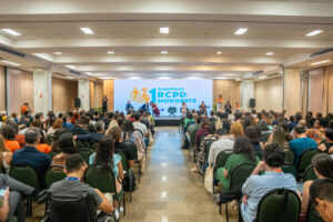 Auditório cheio com muitas pessoas sentadas em fileiras voltadas para um palco ao fundo. O ambiente é interno, amplo, com teto claro e várias luminárias retangulares acesas. As cadeiras são escuras e quase todos os assentos parecem ocupados; há pessoas com diferentes tons de pele e cores de cabelo, muitas usando roupas casuais em cores variadas como verde, azul, laranja e estampadas. Na parte frontal da sala há um palco elevado, com algumas pessoas em pé sobre ele.