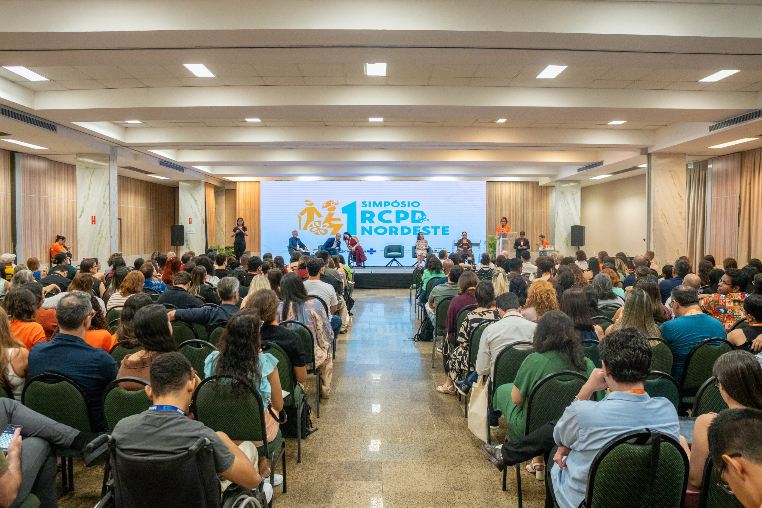 Auditório cheio com muitas pessoas sentadas em fileiras voltadas para um palco ao fundo. O ambiente é interno, amplo, com teto claro e várias luminárias retangulares acesas. As cadeiras são escuras e quase todos os assentos parecem ocupados; há pessoas com diferentes tons de pele e cores de cabelo, muitas usando roupas casuais em cores variadas como verde, azul, laranja e estampadas. Na parte frontal da sala há um palco elevado, com algumas pessoas em pé sobre ele.