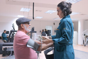Foto em um ambiente amplo e iluminado com equipamentos de fisioterapia e reabilitação ao fundo. Em primeiro plano, uma mulher de pele clara e cabelos pretos curtos, usando jaleco azul petróleo, utiliza um equipamento de aferir pressão em um senhor de cabelos e barba brancos, usando óculos, uma boina e uma camiseta rosa, sentado em frente a ela.