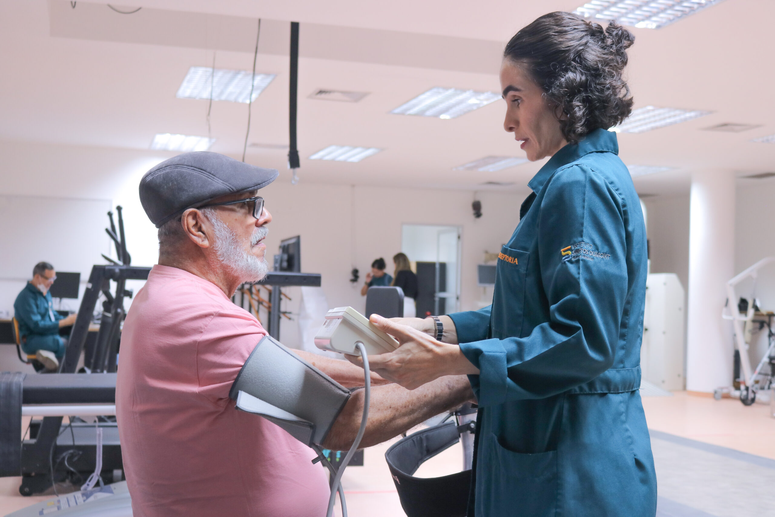 Foto em um ambiente amplo e iluminado com equipamentos de fisioterapia e reabilitação ao fundo. Em primeiro plano, uma mulher de pele clara e cabelos pretos curtos, usando jaleco azul petróleo, utiliza um equipamento de aferir pressão em um senhor de cabelos e barba brancos, usando óculos, uma boina e uma camiseta rosa, sentado em frente a ela.