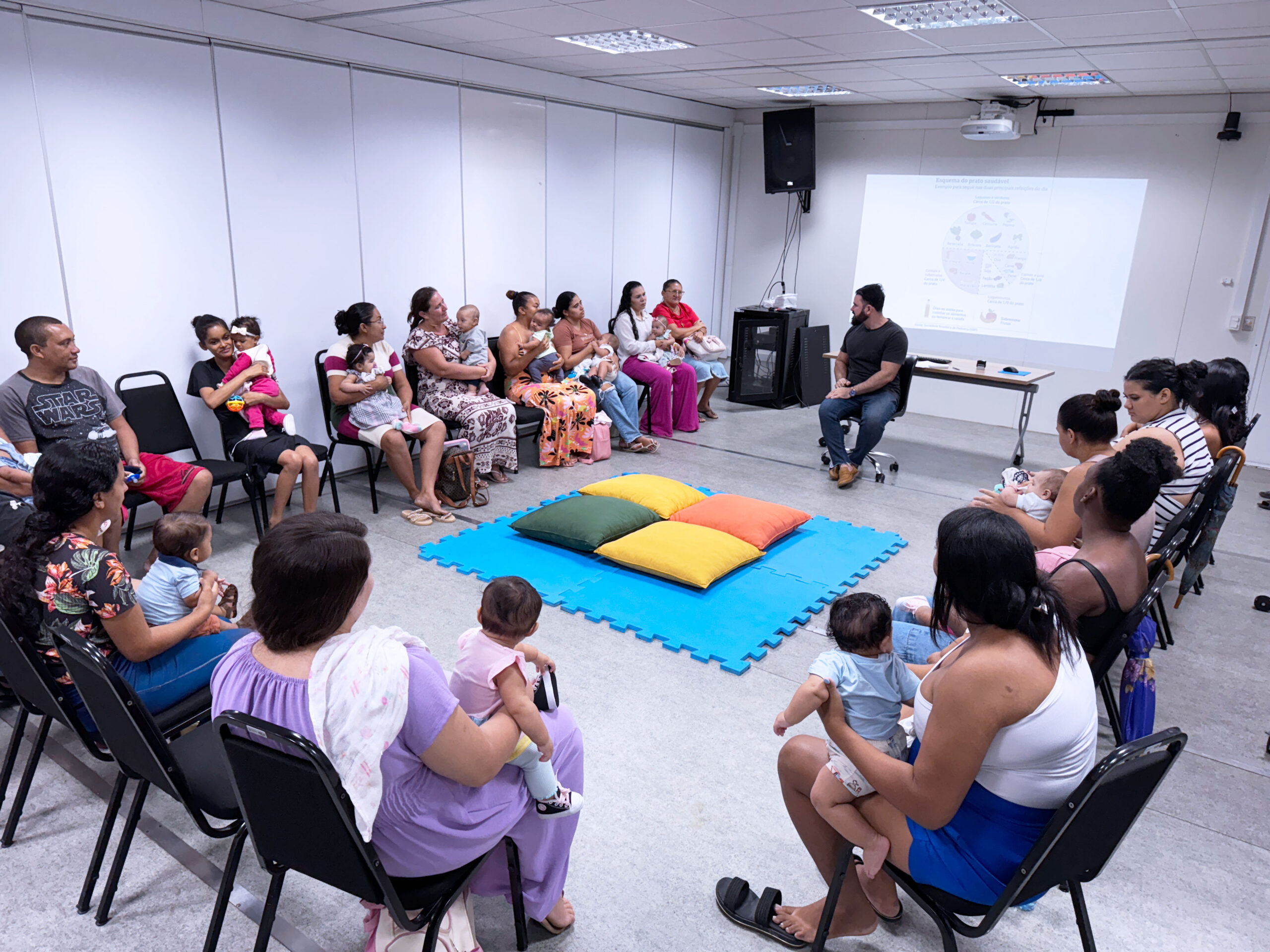 Fotografia colorida em plano médio que captura uma roda de conversa em uma sala de reuniões iluminada. Cerca de quinze adultos, em sua maioria mulheres, estão sentados em cadeiras pretas dispostas em formato de semicírculo. Muitas dessas mulheres seguram bebês de colo nos braços. No centro do semicírculo, sobre o piso claro, há um tapete de EVA azul-celeste com quatro almofadas grandes e quadradas nas cores verde, amarelo e laranja. Ao fundo, um homem veste camiseta preta e calça jeans, sentado em frente a uma tela de projeção branca que exibe o slide de um gráfico circular colorido sobre alimentação saudável, intitulado "Esquema do prato saudável". As pessoas na sala estão atentas, olhando em direção ao palestrante ou conversando entre si. O ambiente possui paredes brancas e divisórias, com caixas de som fixadas no alto. A atmosfera é de um encontro acolhedor e educativo.