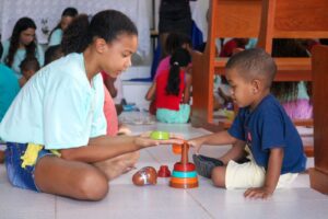 Duas crianças de pele negra, uma menina e um menino, brincam no chão com um brinquedo de encaixar colorido. Ao fundo aparecem outras crianças sentadas e adultos em pé, em um ambiente interno iluminado, com móveis de madeira e várias pessoas interagindo.