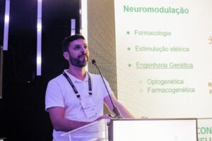 Um homem adulto de pele clara, cabelo curto escuro e barba cheia falando em um púlpito com microfone. Ele está usando camisa polo branca e um crachá pendurado em um cordão preto com inscrições claras. Ao fundo, há um painel com iluminação forte. À direita da foto aparece o slide projetado com o título em verde: “Neuromodulação”, com vários tópicos: “Farmacologia”, “Estimulação elétrica”, “Engenharia Genética”.