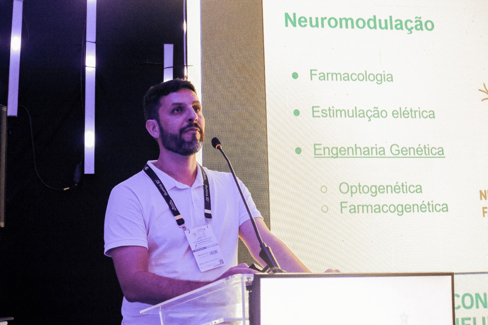 Um homem adulto de pele clara, cabelo curto escuro e barba cheia falando em um púlpito com microfone. Ele está usando camisa polo branca e um crachá pendurado em um cordão preto com inscrições claras. Ao fundo, há um painel com iluminação forte. À direita da foto aparece o slide projetado com o título em verde: “Neuromodulação”, com vários tópicos: “Farmacologia”, “Estimulação elétrica”, “Engenharia Genética”.