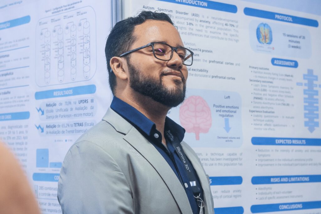 Um homem adulto de barba e cabelo curto escuro, usando óculos de armação fina marrom e um terno cinza-claro com camisa azul-marinho de gola social. Ele está de perfil, levemente sorridente, com expressão tranquila. O fundo é um pôster grande em tons de azul e branco, com muitos blocos de texto, gráficos e ícones, mas o texto está pequeno e desfocado, sem condição de leitura precisa.