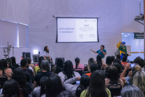 Fotografia de uma palestra em um auditório com paredes brancas texturizadas. Ao fundo, no centro, há uma grande tela de projeção onde se lê o título em letras azuis: "DEFICIÊNCIA É DOENÇA?". Ao lado direito da pergunta, está escrito em destaque: "NÃO! porque representa uma condição permanente de interação entre limitações funcionais e barreiras sociais, e não uma enfermidade a ser 'curada'". Logo abaixo, há uma breve tabela comparativa entre os termos "Doença" (Temporária ou progressiva / tratável) e "Deficiência" (Aspecto da identidade e funcionalidade). Abaixo da tela, duas palestrantes estão de pé: uma à esquerda, com cabelos cacheados e blusa branca, e outra à direita, com cabelos presos, vestindo uma camiseta verde-escura e gesticulando com o braço esquerdo enquanto segura um microfone. Em primeiro plano, vê-se a plateia de costas, composta por dezenas de pessoas sentadas assistindo à apresentação. À direita, ao fundo, há uma divisória de vidro com a inscrição "ESPAÇO NISE / Nise da Silveira" e um manequim vestindo uma camisa verde e amarela com o número 10.