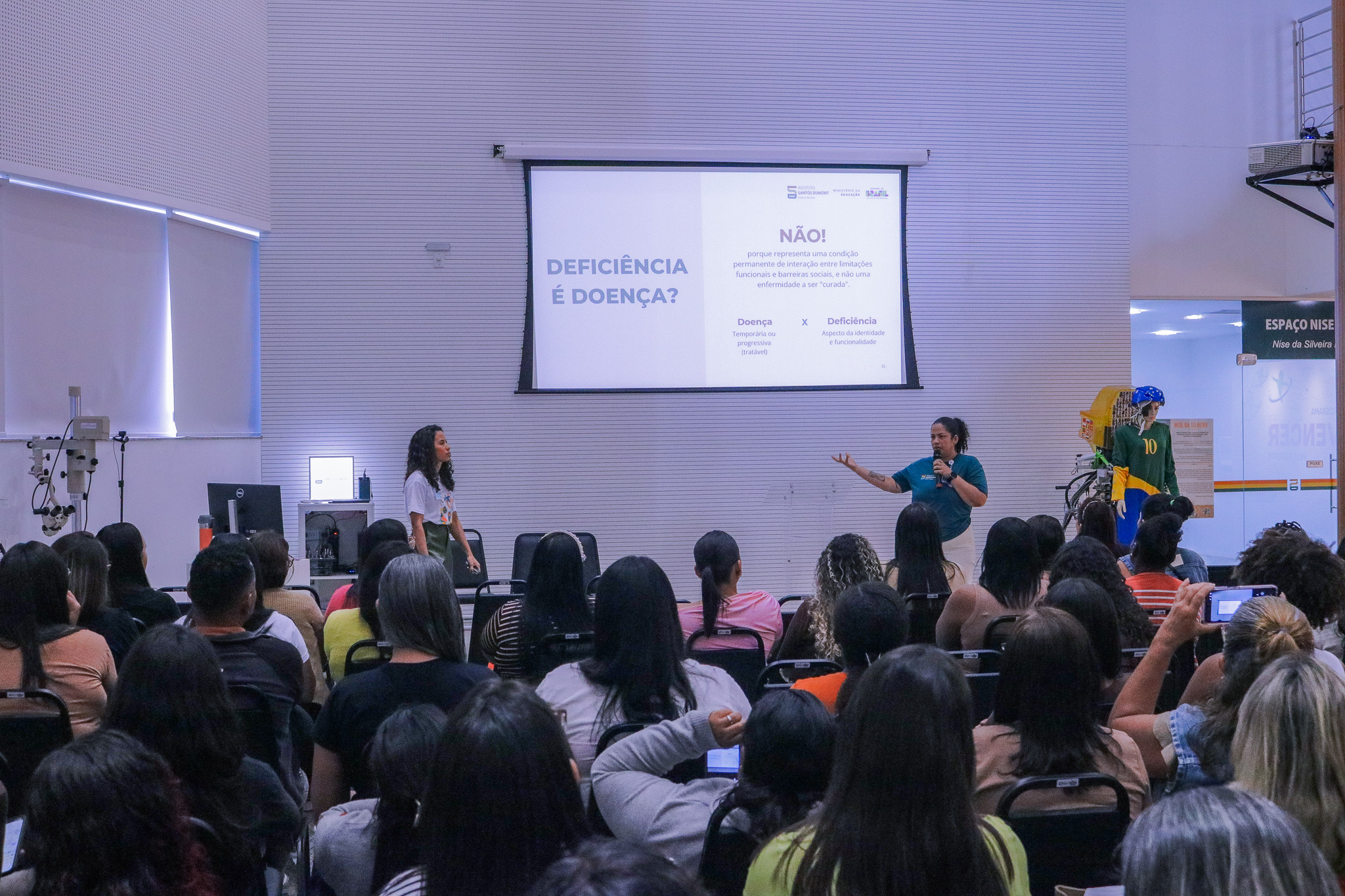 Fotografia de uma palestra em um auditório com paredes brancas texturizadas. Ao fundo, no centro, há uma grande tela de projeção onde se lê o título em letras azuis: "DEFICIÊNCIA É DOENÇA?". Ao lado direito da pergunta, está escrito em destaque: "NÃO! porque representa uma condição permanente de interação entre limitações funcionais e barreiras sociais, e não uma enfermidade a ser 'curada'". Logo abaixo, há uma breve tabela comparativa entre os termos "Doença" (Temporária ou progressiva / tratável) e "Deficiência" (Aspecto da identidade e funcionalidade). Abaixo da tela, duas palestrantes estão de pé: uma à esquerda, com cabelos cacheados e blusa branca, e outra à direita, com cabelos presos, vestindo uma camiseta verde-escura e gesticulando com o braço esquerdo enquanto segura um microfone. Em primeiro plano, vê-se a plateia de costas, composta por dezenas de pessoas sentadas assistindo à apresentação. À direita, ao fundo, há uma divisória de vidro com a inscrição "ESPAÇO NISE / Nise da Silveira" e um manequim vestindo uma camisa verde e amarela com o número 10.