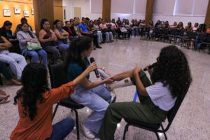 Fotografia em plano aberto de um auditório onde ocorre uma dinâmica prática. Ao centro, duas mulheres estão sentadas em cadeiras pretas, de perfil uma para a outra. A mulher da esquerda, com cabelos escuros presos e usando uma camiseta verde-escura e calça jeans clara, segura um microfone na mão esquerda e, com a mão direita, examina ou demonstra algo na perna de outra mulher. Esta segunda mulher está à direita, veste camiseta branca e calça verde-oliva, e está com a perna esquerda estendida, calçando uma bota ortopédica branca (estilo Robofoot). Uma terceira mulher, de blusa laranja e calça jeans, está ajoelhada em primeiro plano à esquerda, com o braço estendido em direção à dinâmica. Ao fundo, uma grande plateia composta majoritariamente por mulheres está sentada em semicírculo em cadeiras pretas, prestando atenção na demonstração. O chão do local é de granito polido e as paredes têm painéis de madeira clara.