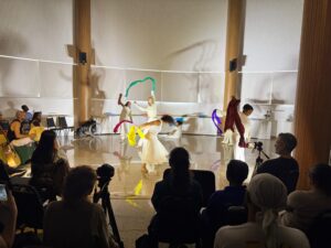 Apresentação artística em um espaço interno. No centro, há pessoas em trajes claros, parecidos com roupas de performance ou dança, em movimento, balançando e girando tecidos coloridos. À frente e ao redor deles, muitas pessoas aparecem apenas como silhuetas escuras, sentadas e de pé, formando o público que acompanha a apresentação. Entre elas, há ao menos uma câmera montada em tripé apontada para o palco, registrando o evento.