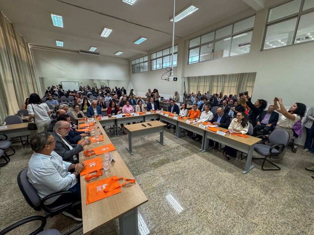 Fotografia em plano aberto e ângulo elevado mostrando uma reunião institucional em uma grande sala de conferências com mesas dispostas em formato de "U". Ao redor das mesas de madeira clara, dezenas de homens e mulheres estão sentados em cadeiras executivas cinzas, acompanhando a reunião. Sobre as mesas há copos de água, microfones, placas de identificação e sacolas promocionais de cor laranja com a marca "PAX". À direita, uma mulher em pé registra o evento tirando uma foto com o celular. Ao fundo, um espelho amplo reflete o restante da sala e da plateia acomodada em fileiras de cadeiras.