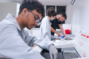 Fotografia de rotina de trabalho científico em laboratório. Em primeiro plano à esquerda, um jovem pesquisador de cabelo cacheado, óculos e jaleco cinza claro com luvas descartáveis opera com atenção um equipamento laboratorial (criostato Leica). Ao fundo, em segundo plano levemente desfocado, uma pesquisadora também de jaleco e um colega de camiseta preta trabalham juntos em uma bancada examinando documentos ou amostras.