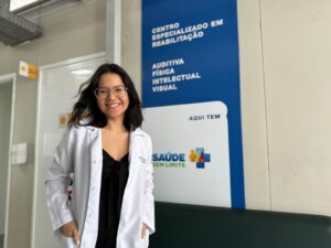 A imagem apresenta uma mulher jovem, sorridente, posando em um ambiente institucional de saúde. Ela tem pele clara, cabelos pretos na altura dos ombros e usa óculos de grau com armação arredondada. Ela veste uma blusa preta com um nó no decote e um jaleco branco aberto por cima, com as mãos apoiadas nos bolsos do jaleco. Ao fundo, há uma parede clara e uma placa vertical azul e branca fixada nela. Texto contido na placa (verbatim): CENTRO ESPECIALIZADO EM REABILITAÇÃO AUDITIVA FÍSICA INTELECTUAL VISUAL AQUI TEM SAÚDE SEM LIMITE