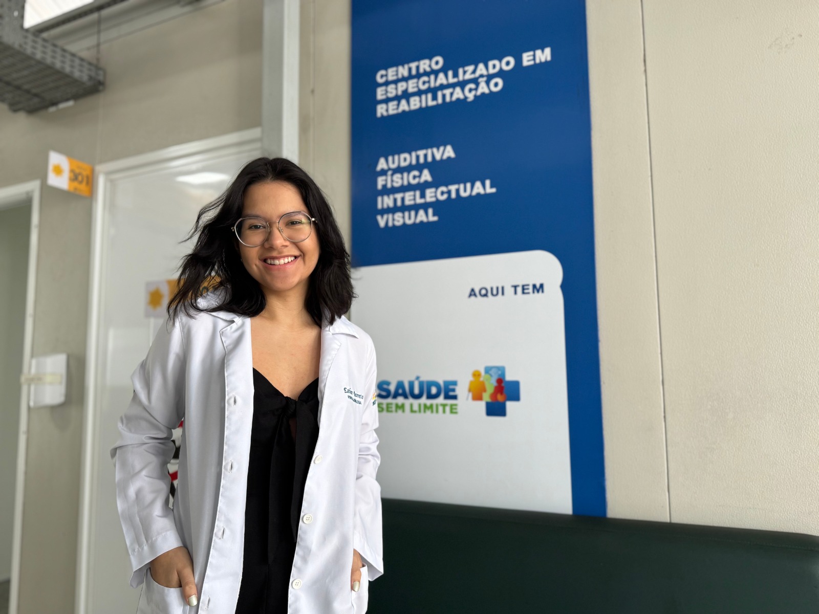 A imagem apresenta uma mulher jovem, sorridente, posando em um ambiente institucional de saúde. Ela tem pele clara, cabelos pretos na altura dos ombros e usa óculos de grau com armação arredondada. Ela veste uma blusa preta com um nó no decote e um jaleco branco aberto por cima, com as mãos apoiadas nos bolsos do jaleco. Ao fundo, há uma parede clara e uma placa vertical azul e branca fixada nela. Texto contido na placa (verbatim): CENTRO ESPECIALIZADO EM REABILITAÇÃO AUDITIVA FÍSICA INTELECTUAL VISUAL AQUI TEM SAÚDE SEM LIMITE