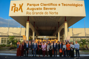 Fotografia externa em plano geral de um grande grupo de homens e mulheres reunidos para uma foto oficial em frente à fachada do Parque Científico e Tecnológico Augusto Severo. A estrutura do portal do prédio possui uma grande marquise branca com letras na cor amarela onde se lê: "PAX - Parque Científico e Tecnológico Augusto Severo - Rio Grande do Norte". O grupo com cerca de 30 pessoas está posicionado no centro, sorrindo em direção à câmera, sob iluminação natural do dia.