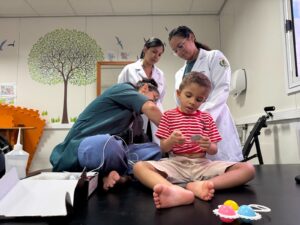 Fotografia de atendimento e avaliação clínica multidisciplinar em saúde infantil. Em primeiro plano, uma criança pequena de camiseta listrada em vermelho e branco e bermuda bege está sentada descalça sobre uma mesa de exame, manipulando um pequeno objeto entre as mãos. Ao seu redor, três profissionais de saúde realizam o acompanhamento: À esquerda, uma profissional de uniforme verde-escuro está curvada realizando procedimentos técnicos ou posicionando sensores/fios conectados a um equipamento. Ao fundo e à direita, duas residentes vestindo jalecos brancos observam atentamente a criança e o procedimento realizado. O ambiente é decorado com temática infantil, apresentando um mural com desenho de árvore e pássaros na parede ao fundo.