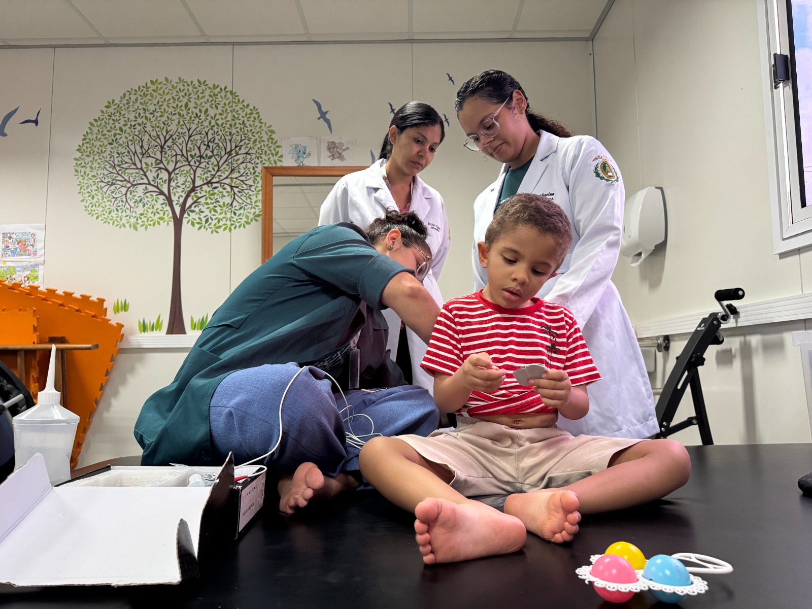 Fotografia de atendimento e avaliação clínica multidisciplinar em saúde infantil. Em primeiro plano, uma criança pequena de camiseta listrada em vermelho e branco e bermuda bege está sentada descalça sobre uma mesa de exame, manipulando um pequeno objeto entre as mãos. Ao seu redor, três profissionais de saúde realizam o acompanhamento: À esquerda, uma profissional de uniforme verde-escuro está curvada realizando procedimentos técnicos ou posicionando sensores/fios conectados a um equipamento. Ao fundo e à direita, duas residentes vestindo jalecos brancos observam atentamente a criança e o procedimento realizado. O ambiente é decorado com temática infantil, apresentando um mural com desenho de árvore e pássaros na parede ao fundo.