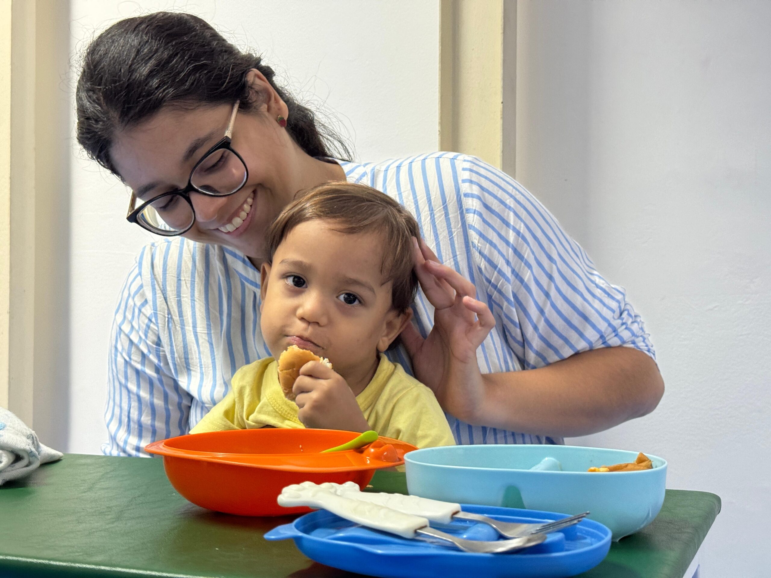 Fotografia de refeição e apoio no desenvolvimento infantil. Em plano médio e com enquadramento horizontal, uma mulher e uma criança pequena estão acomodadas em uma mesa de tampo verde: Mulher: À esquerda, vestindo blusa listrada em azul e branco e usando óculos de armação escura, sorri carinhosamente enquanto apoia a mão suavemente na cabeça da criança. Criança: Ao centro, vestindo blusa amarela, olha para a frente enquanto leva um alimento (pão ou bolinho) à boca. Mesa: Em primeiro plano, estão dispostos recipientes plásticos de refeição — uma tigela alaranjada com colher verde, um prato azul-claro e uma tampa/suporte azul com talheres infantis.